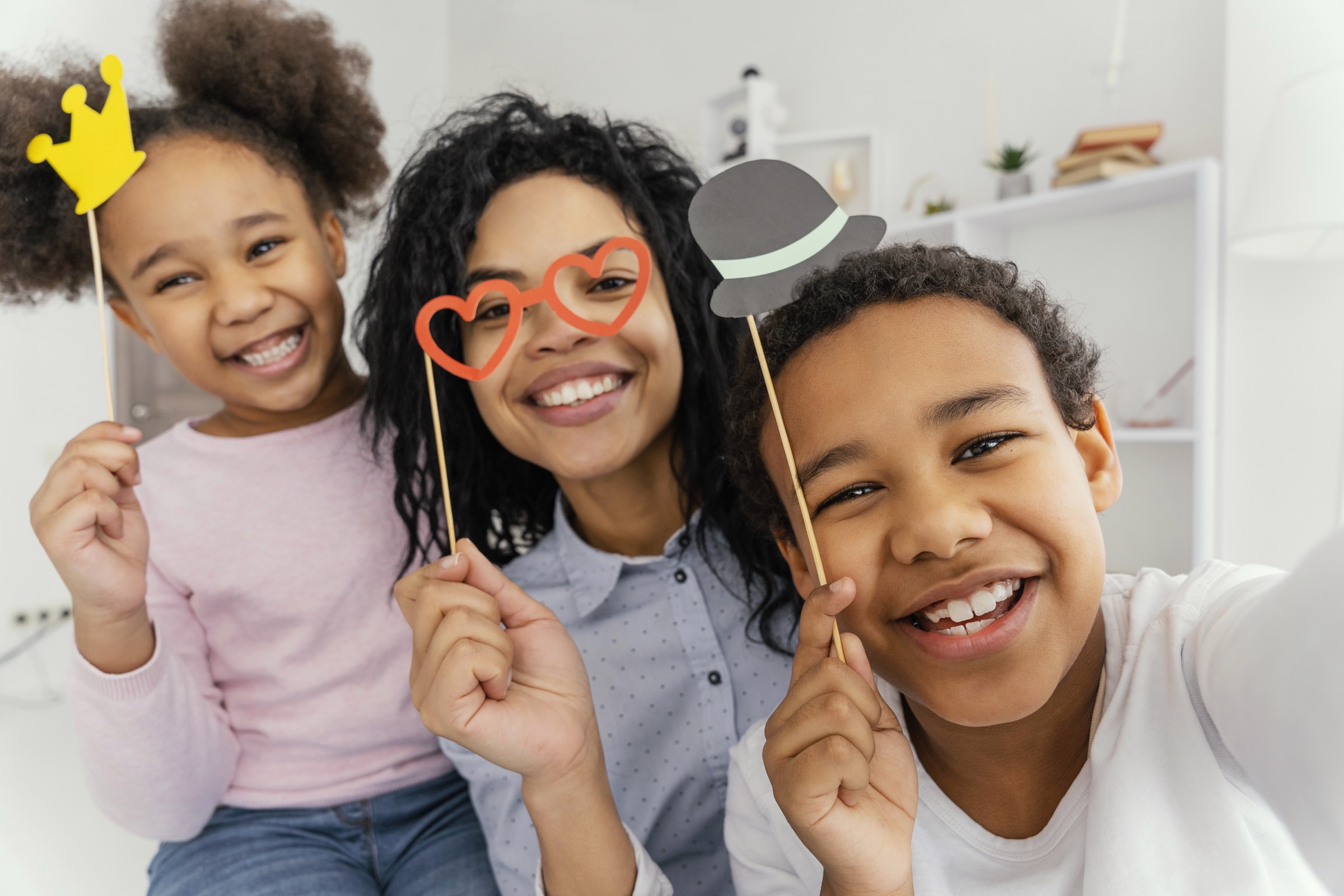 A mother and her two children laughing into a selfie, holding paper props: a gold crown, red heart glasses and a grey hat